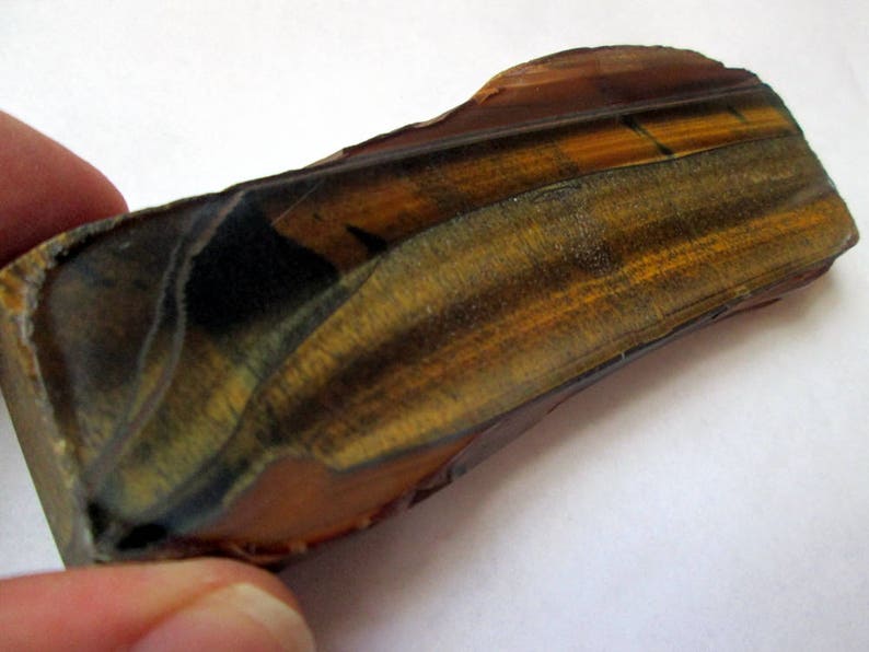 May include: A raw, uncut piece of tiger's eye gemstone with a brown, gold, and black color pattern. The stone has a smooth, polished surface and a slightly curved shape.