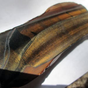May include: A large, raw, brown and gold tiger's eye gemstone with a unique, banded pattern. The stone has a smooth, polished surface and is a natural, raw specimen.