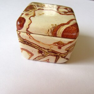 May include: A small, square box made of brown, white, and red marbled stone. The box has a hinged lid and is closed.