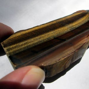 May include: A polished, rectangular piece of tiger's eye gemstone with brown, gold, and black stripes. The stone has a smooth, glossy finish.