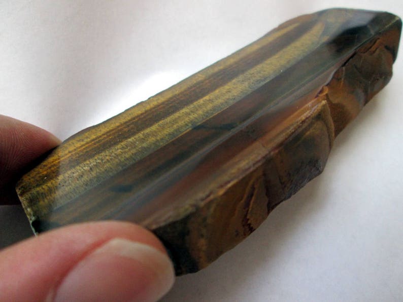 May include: A raw, unpolished piece of tiger's eye gemstone with a natural, irregular shape. The stone has a rich brown, gold, and black color pattern with a glossy finish.