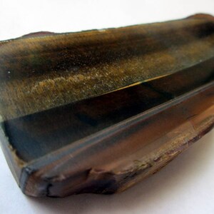 May include: A raw, unpolished piece of tiger's eye gemstone with a brown, gold, and black color pattern. The stone has a rough, natural texture.