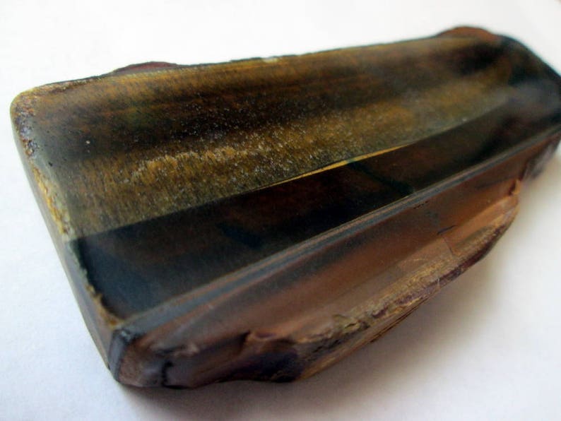 May include: A raw, unpolished piece of tiger's eye gemstone with a brown, gold, and black color pattern. The stone has a rough, natural texture.