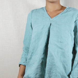 Könnte beinhalten: Ein hellblaues, langärmeliges Leinenhemd mit V-Ausschnitt und einer gefalteten Vorderseite. Das Hemd wird mit einer blauen Jeans getragen.