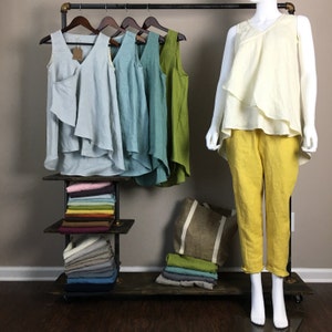 May include: A display of women's linen clothing in various colors. Tank tops in light blue, teal, and green hang on a rack. A mannequin wears a cream-colored top and yellow pants. Folded fabric and a tote bag are also displayed.
