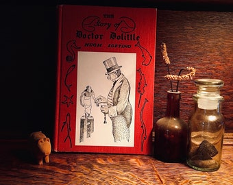 Puede incluir: Un libro de tapa dura rojo titulado "The Story of Doctor Dolittle" de Hugh Lofting. La portada presenta una ilustración en blanco y negro de un hombre con un sombrero de copa y un mono.
