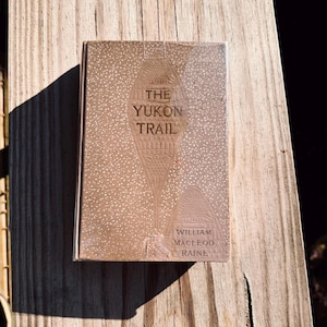 May include: A vintage book titled "The Yukon Trail" by William MacLeod Raine. The cover is a light brown with a detailed illustration of a snowshoe and a cabin. The title is in a bold, serif font. The book is resting on a wooden surface.
