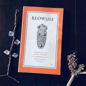 Puede incluir: Un libro de bolsillo naranja con el título "BEOWULF" impreso en negro. El libro es un clásico de Penguin y fue traducido por David Wright. La portada presenta una ilustración en blanco y negro de un objeto metálico decorativo.