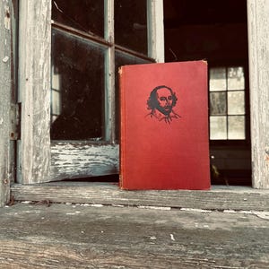 Puede incluir: Un libro de tapa dura rojo con una ilustración en blanco y negro de William Shakespeare en la portada. El libro está sentado en un alféizar de la ventana.
