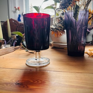 May include: A dark red glass goblet with a clear stem and base sits on a wooden table. The goblet is cylindrical with a flared rim. Dried flowers in a purple vase are in the background.