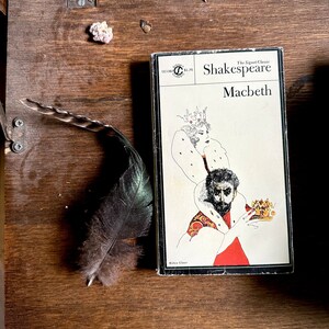 May include: A vintage paperback copy of Shakespeare's Macbeth with a black and white illustration of a king and queen on the cover. The book is on a wooden surface with a feather and a small white object.