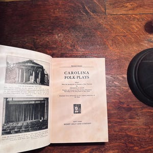 May include: An open book titled "CAROLINA FOLK-PLAYS" with black and white images of a theatre. The book is open on a wooden surface. The text includes "SECOND SERIES" and "HENRY HOLT AND COMPANY".