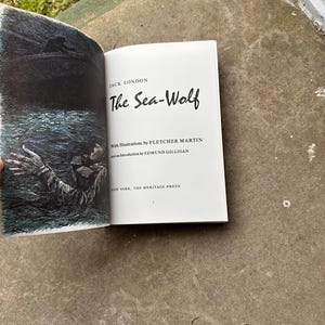 May include: An open book titled "The Sea-Wolf" by Jack London. The book's cover features a dark illustration of a person in the water. The title is in a bold, stylized font. The book is from The Heritage Press, New York.
