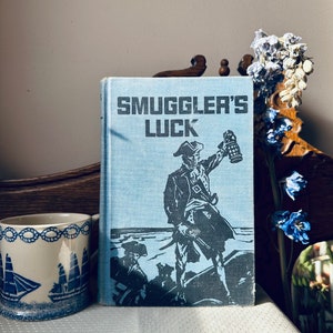 Op de afbeelding: Een vintage blauw boek met de titel "Smuggler's Luck" op de omslag. De omslag toont een zwart-witte illustratie van een piraat die een lantaarn en een zwaard vasthoudt.