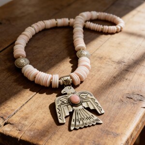 May include: A necklace featuring pale pink disc-shaped beads interspersed with small metallic spacers and two decorative round silver beads. A large silver-toned bird pendant with engraved wing details and a central oval pink stone hangs from the center.