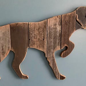 May include: A wall-mounted wooden cutout shaped like a pointing dog, constructed from vertical planks of weathered brown wood with a visible grain. In the bottom right corner, a black logo features a dog silhouette and the text PAW-LICK Design.