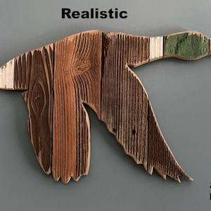May include: Wooden duck wall art in flight, crafted from various shades of brown wood with a green and white head. The word "Realistic" is above the duck. The design is by Paw-Lick Design.
