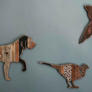 May include: Wooden wall art featuring a dog, a flying bird, and a pheasant. The dog is walking, the bird is in flight, and the pheasant is standing. The art is made of various wood tones and textures. The logo "PAW-LICK Design" is in the upper left corner.