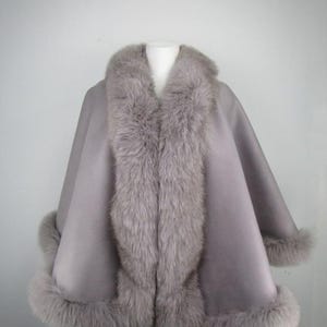 May include: A grey cape with a faux fur collar and trim. The cape has a wide collar and trim around the sleeves and hem. The garment is displayed on a mannequin against a plain background.