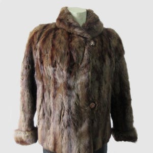 May include: A brown fur jacket with a rounded collar and two buttons. The jacket has long sleeves with fur cuffs. The garment is displayed on a mannequin against a white background. The fur appears to be a mix of brown and reddish-brown tones.