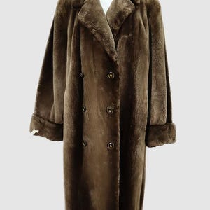 May include: A long, brown faux fur coat with a double-breasted closure and notched lapels. The coat features long sleeves with turned-up cuffs and gold-colored buttons. The coat is displayed on a white mannequin.