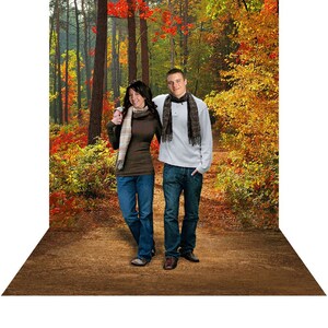 May include: A couple walks along a path in a forest with fall foliage. The woman is wearing a brown sweater and a plaid scarf. The man is wearing a white sweater and a brown scarf.