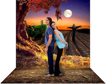 May include: A couple stands in a field with a tractor, a scarecrow, and a full moon in the background. The man is wearing a blue shirt and jeans, and the woman is wearing a blue shirt and black pants. The field is covered in dry grass and dirt.