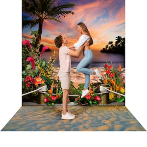 May include: A couple is embracing on a tropical beach. The man is holding the woman in the air as they look at each other and smile. The background is a sunset over the ocean with palm trees and lush foliage.