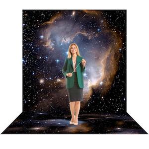 May include: A person wearing a dark green blazer, a light pink top, and a dark skirt, holding a thin white conductor baton. The background features a vibrant nebula with swirling orange and blue gases and numerous bright stars in deep space.