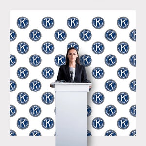 Puede incluir: Una mujer con un traje negro se encuentra detrás de un podio blanco con un micrófono. El fondo es un telón de fondo blanco con un patrón repetitivo de círculos azules y blancos con el texto "Kiwanis International" en su interior.
