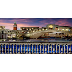 Puede incluir: Un puente blanco con arcos sobre una masa de agua al atardecer. El puente est&aacute; iluminado con luces y hay edificios a ambos lados del puente. La escena est&aacute; enmarcada por una barandilla de piedra.