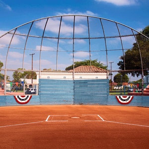May include: A baseball field with a blue wooden backstop and a chain link fence. The field is surrounded by trees and a building. There are two American flags hanging on the backstop.