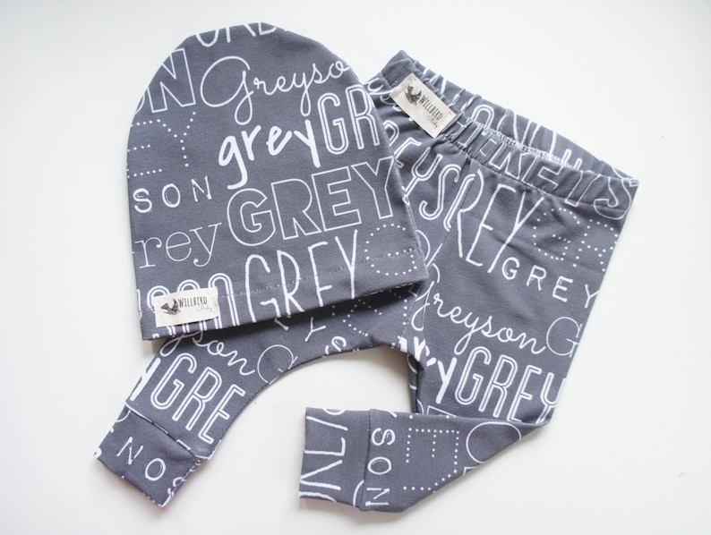 K&ouml;nnte beinhalten: Graues und wei&szlig;es Baby-Leggings- und M&uuml;tzen-Set mit dem Wort "Grey", das in verschiedenen Schriftarten und Gr&ouml;&szlig;en wiederholt wird.
