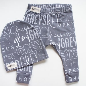 K&ouml;nnte beinhalten: Graues und wei&szlig;es Baby-Leggings- und M&uuml;tzen-Set mit einem sich wiederholenden Muster des Wortes "Grey" in verschiedenen Schriftarten und Gr&ouml;&szlig;en.