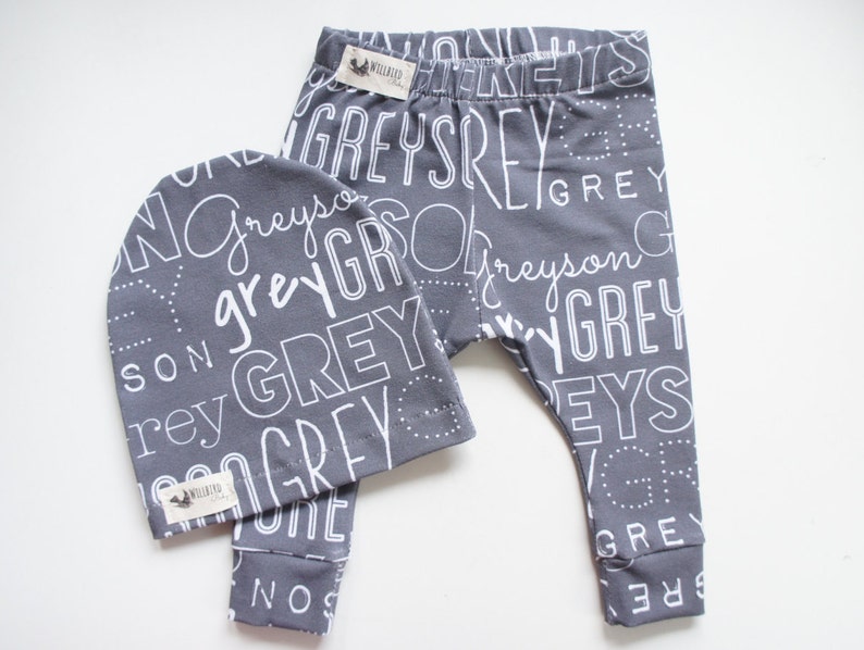 K&ouml;nnte beinhalten: Graues und wei&szlig;es Baby-Leggings- und M&uuml;tzen-Set mit einem sich wiederholenden Muster des Wortes "Grey" in verschiedenen Schriftarten und Gr&ouml;&szlig;en.