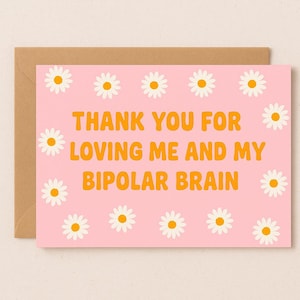 May include: A pink greeting card with the text "THANK YOU FOR LOVING ME AND MY BIPOLAR BRAIN" in orange, surrounded by daisy illustrations. The card is inside a brown envelope.