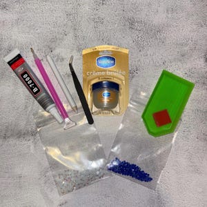 May include: A collection of craft supplies including a tube of B-7000 glue, a jar of Vaseline crème brûlée lip therapy jelly, tweezers, and various tools. Two small bags contain clear and blue rhinestones. A green tray holds a red square.
