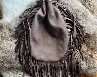 May include: A brown leather bag with a drawstring closure and fringe detailing. The bag is made of soft, supple leather and has a rustic, vintage look.