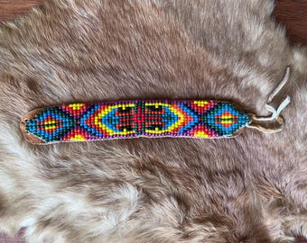 May include: A colorful beaded bracelet with a geometric pattern in shades of red, yellow, blue, green, and purple. The bracelet is made of leather and has a simple tie closure.