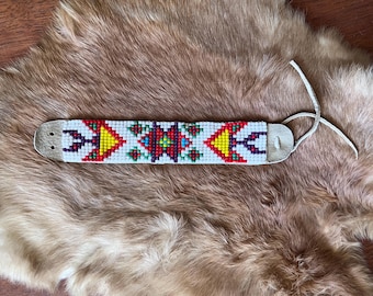 May include: A white leather bracelet with a colorful beaded design. The design features geometric shapes in red, yellow, blue, green, and purple.