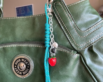 Könnte beinhalten: Eine grüne Ledertasche mit einem silbernen Reißverschluss und einem blauen und weißen Seil-Schlüsselanhänger mit einem roten Erdbeer-Anhänger. Die Tasche trägt den Schriftzug "Nine West" auf der Vorderseite.