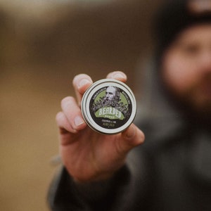 Peut inclure: Une boîte ronde de baume à barbe avec une illustration en noir et blanc d'un homme barbu. La boîte est étiquetée "Bearded Nomad Cedarwood & Lime Beard Balm".