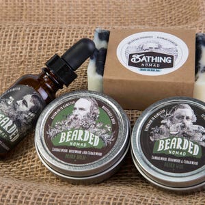 Op de afbeelding: Een set baardverzorgingsproducten in een rustieke omgeving. De set bevat een fles baardolie, twee blikken baardbalsem en een zeep. De producten zijn geëtiketteerd met "Bearded Nomad" en hebben een geur van sandelhout, rozenhout en cederhout.