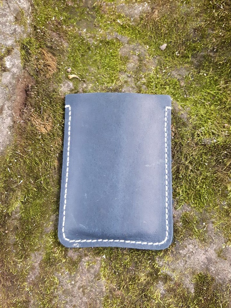 May include: A dark blue leather pouch with white stitching. The pouch is lying on a bed of green moss.
