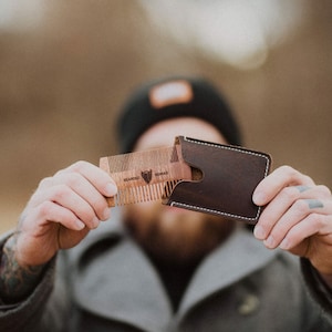May include: A brown leather wallet with a stitched edge and a wooden comb with the text "Bearded Nomad" inside. The comb is partially pulled out of the wallet.