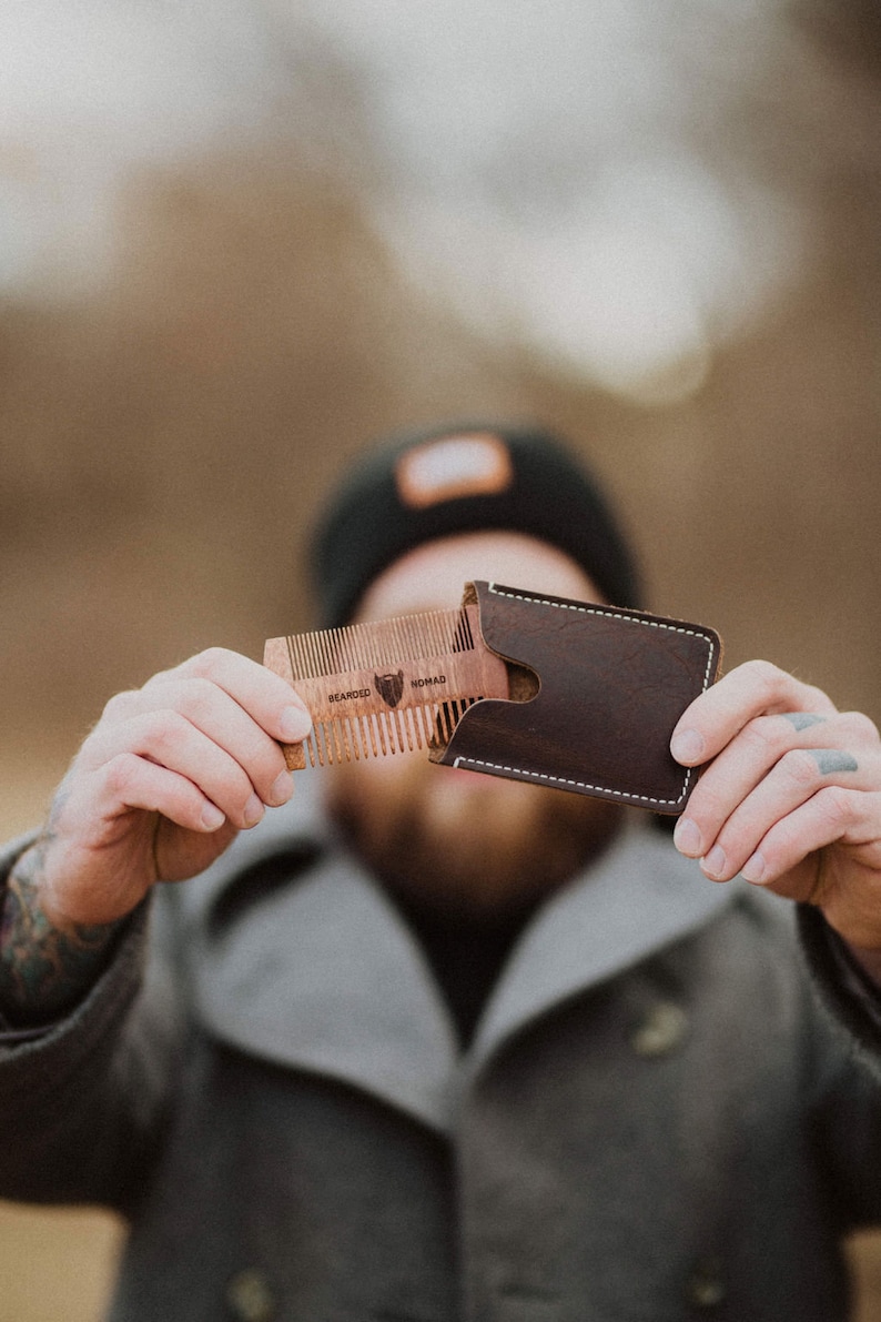 May include: A brown leather wallet with a stitched edge and a wooden comb with the text "Bearded Nomad" inside. The comb is partially pulled out of the wallet.