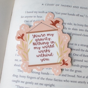 K&ouml;nnte beinhalten: Ein dekorativer Aufkleber mit einem rosa Rahmen und dem Text "you're my gravity. Nothing in my world works without you." Der Aufkleber hat ein florales Design und Fledermaus-Silhouetten. Er liegt auf einem offenen Buch.