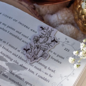 May include: A close-up of an open book with a bookmark featuring a black and white illustration of a sword and flowers. The book is titled "A Court of Thorns and Roses" and the page number is 223. The book is resting on a white surface with a golden decorative object and white flowers.