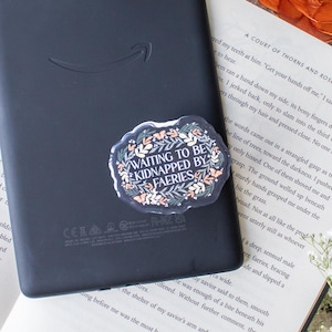 May include: A black e-reader with a sticker that reads "Waiting to be Kidnapped by Faeries" is placed on an open book. The scene is decorated with dried orange slices, white flowers, and a gold-colored decorative sphere.