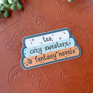 May include: A sticker with a blue, white, and light blue color scheme. The sticker has the text "tea, cozy sweaters, & fantasy novels" with stars around the text.